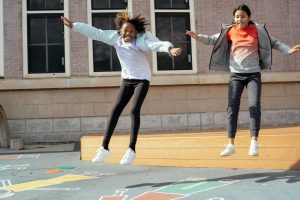 Full body of happy multiracial girls jumping off wooden scene onto playground while spending free time after lessons on backyard of school on sunny day